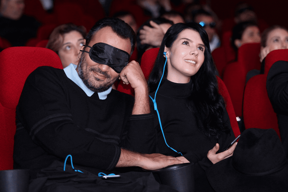 ผู้ชายและผู้หญิงนั่งชมภาพยนตร์พร้อมใส่หูฟัง โดยผู้ชายมีผ้าปิดตาพร้อมรอยยิ้มของทั้งคู่